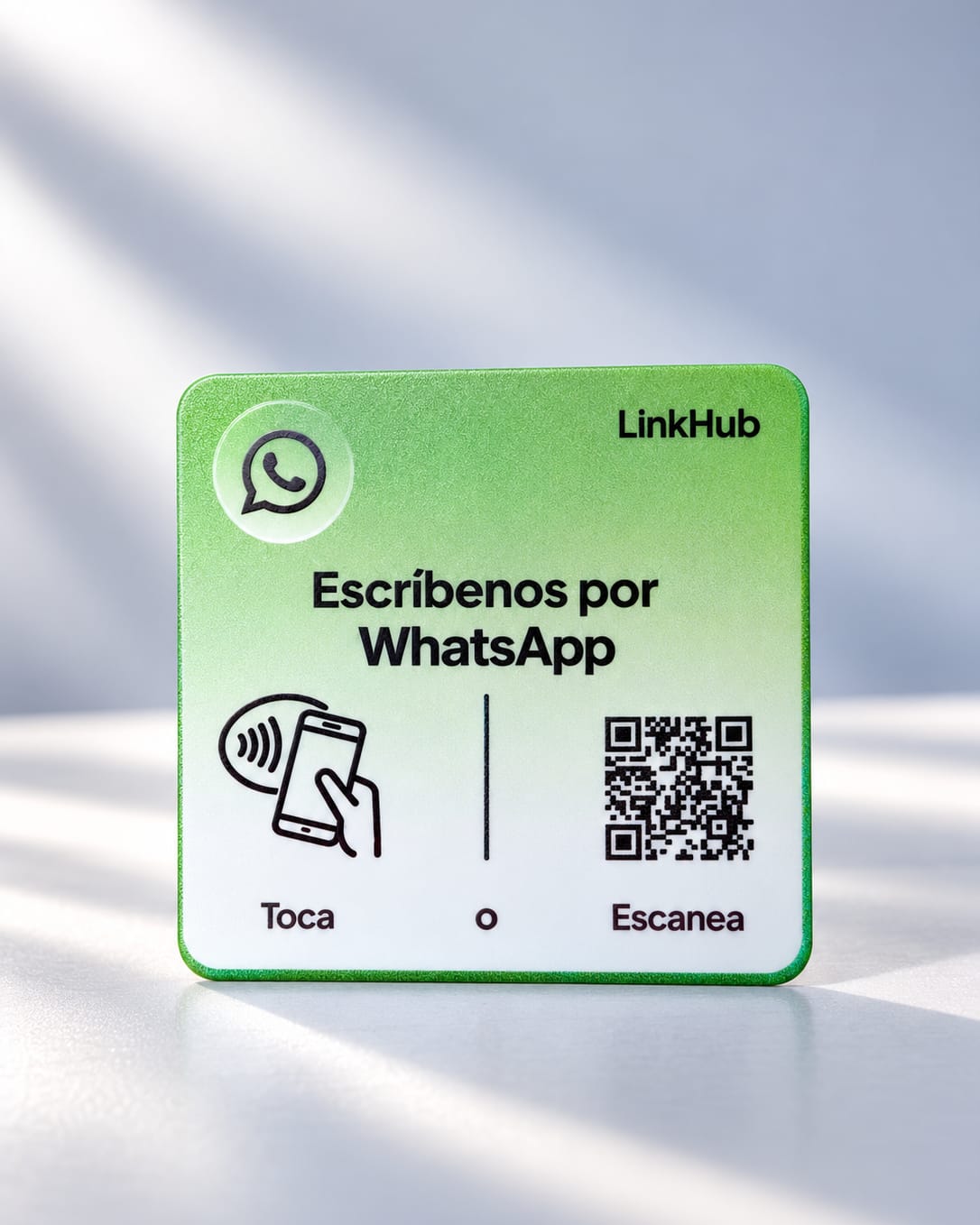 Placa mini verde de LinkHub con «Escríbenos por WhatsApp», el ícono para tocar y su código QR, sobre una superficie clara con franjas de luz