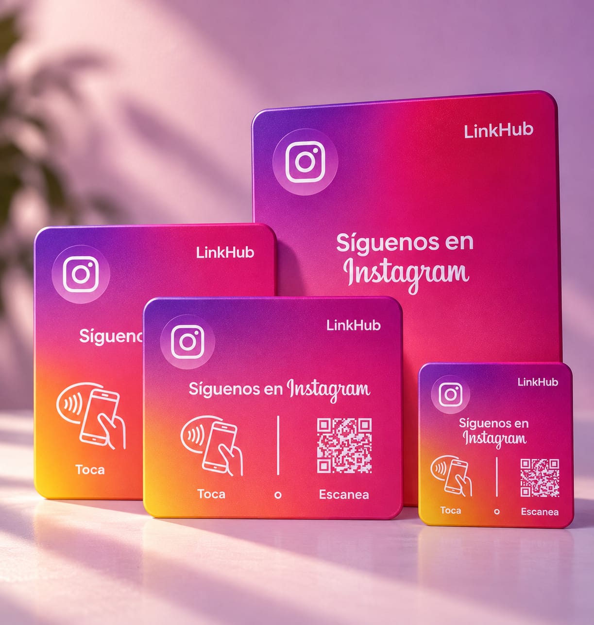 Cuatro placas de LinkHub de distintos tamaños, de la mini a la de mesa, en degradado morado, rosado y naranja, con un ícono de cámara, sobre una mesa con luz de tarde