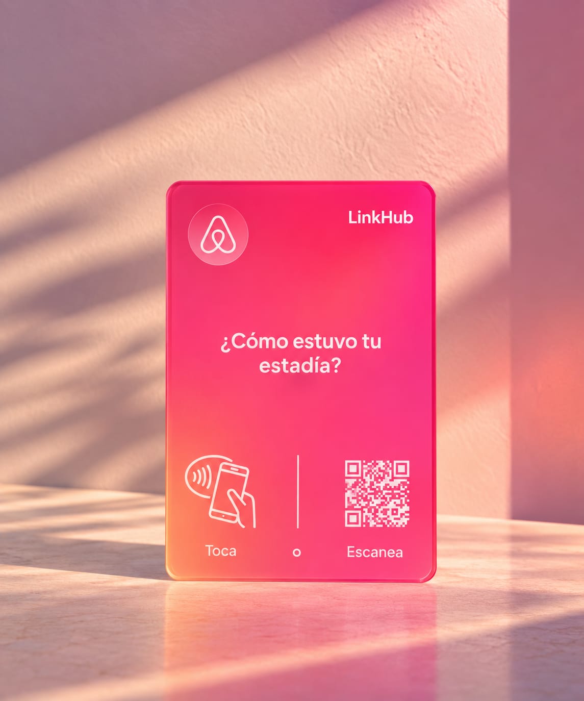 Placa de LinkHub en degradado rosado y naranja con el logo de Airbnb, el ícono para tocar y un código QR, sobre una mesa con la luz de la tarde entrando por la ventana