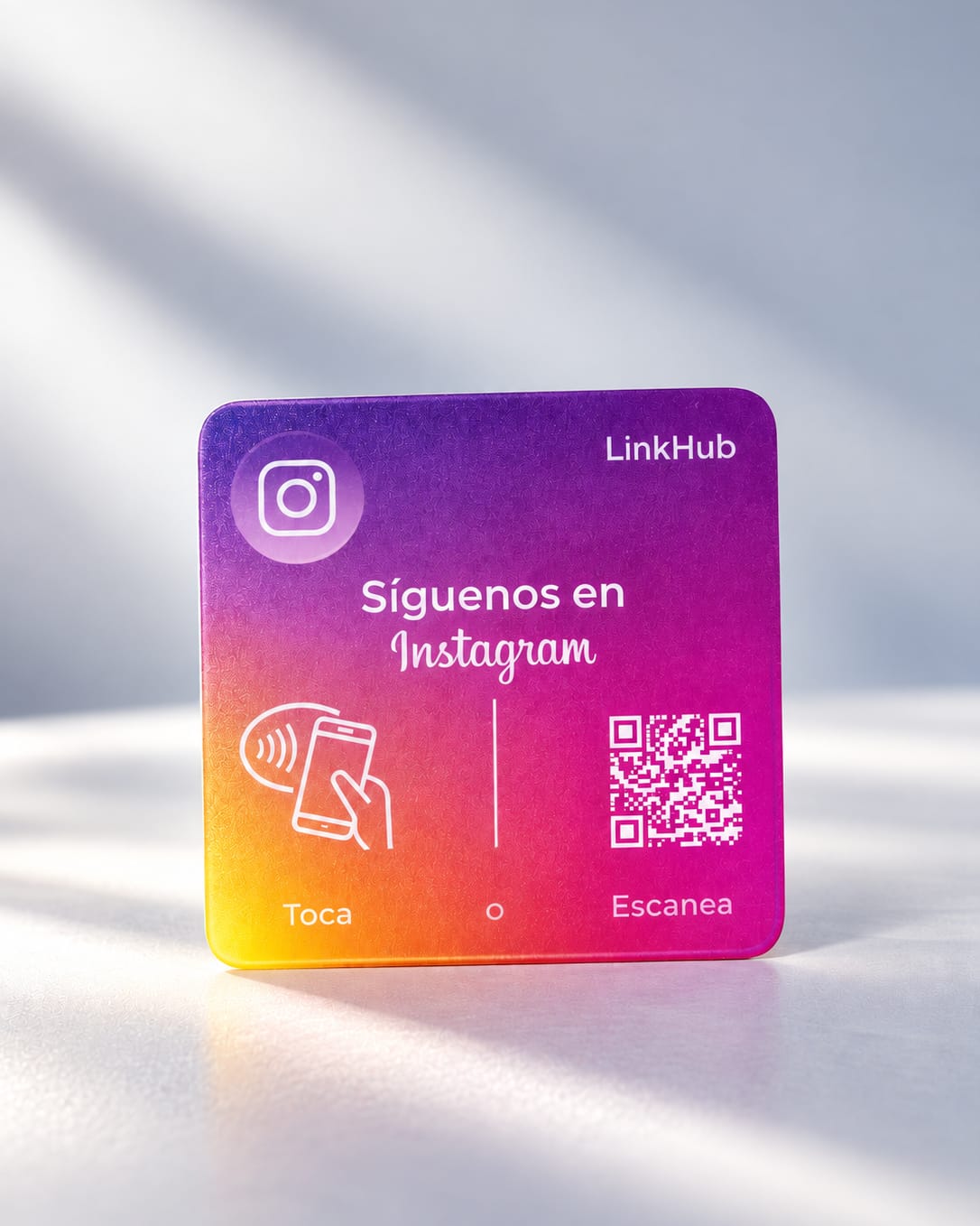 Placa mini de LinkHub con degradado morado, rosado y amarillo, el logo de una cámara, un texto para seguir al negocio en redes y un código QR en blanco, con luz de ventana